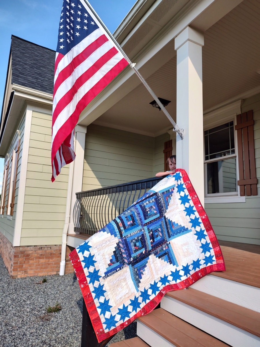 One of Bea's Children Holding the Finished Scrappy Patriotic Log Cabin Star Quilt On Their Front Porch