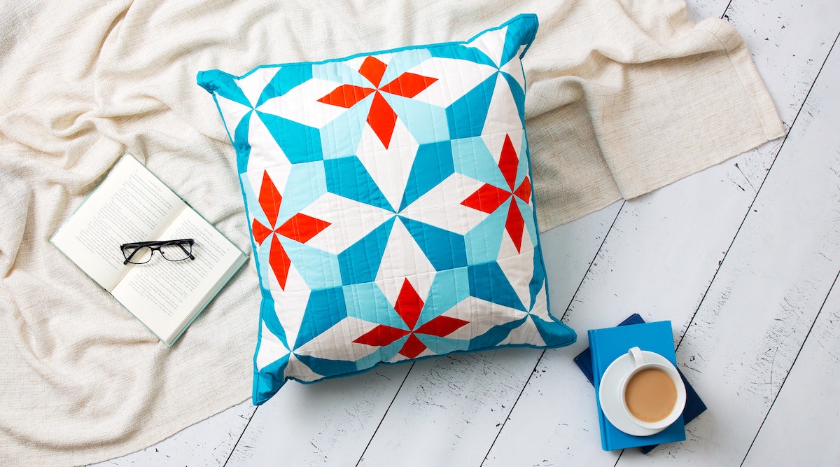 Red, teal and white Hattie's Choice pattern quilted pillow on a white wood floor next to coffee, a blanket, a book and glasses.