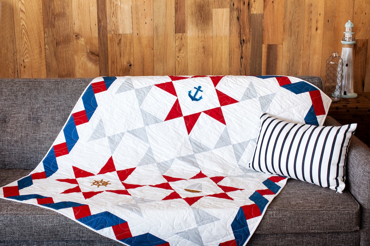 pieced red, white, grey and blue quilt with nautical applique shapes on grey couch with wooden background