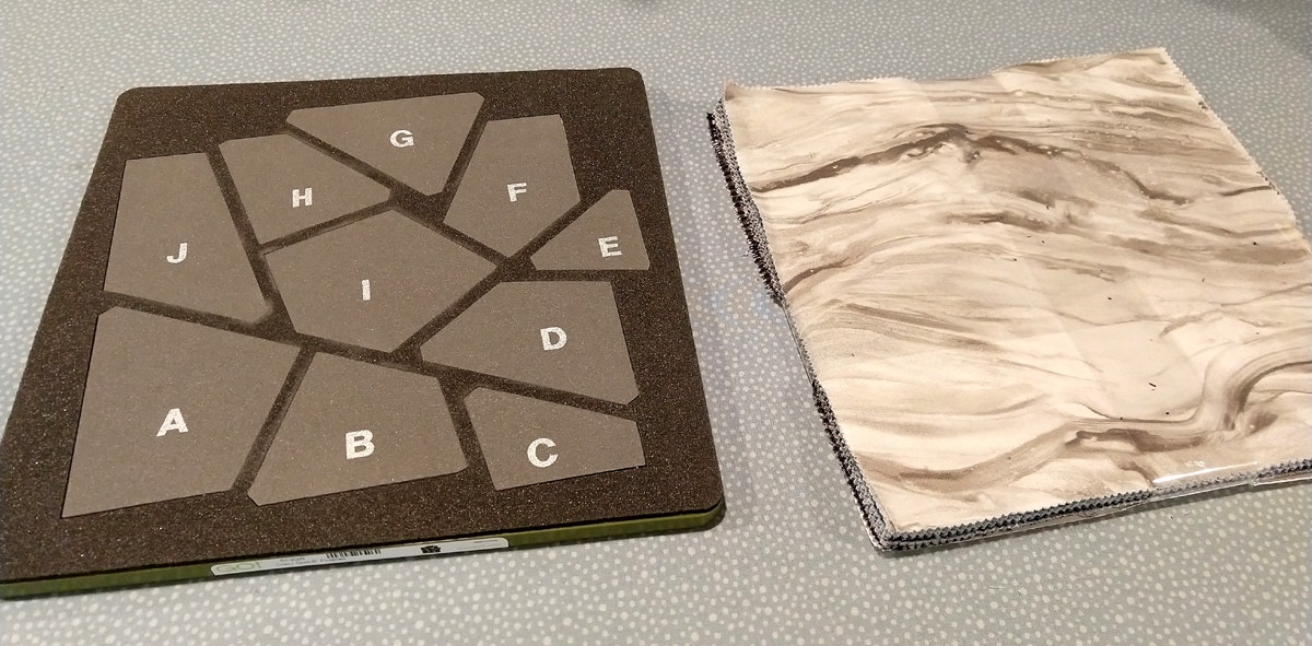 AccuQuilt GO Crazy Quilt 6-inch finished quilt block die next to Michael Millers Fabric Strata layer cake on a grey table.